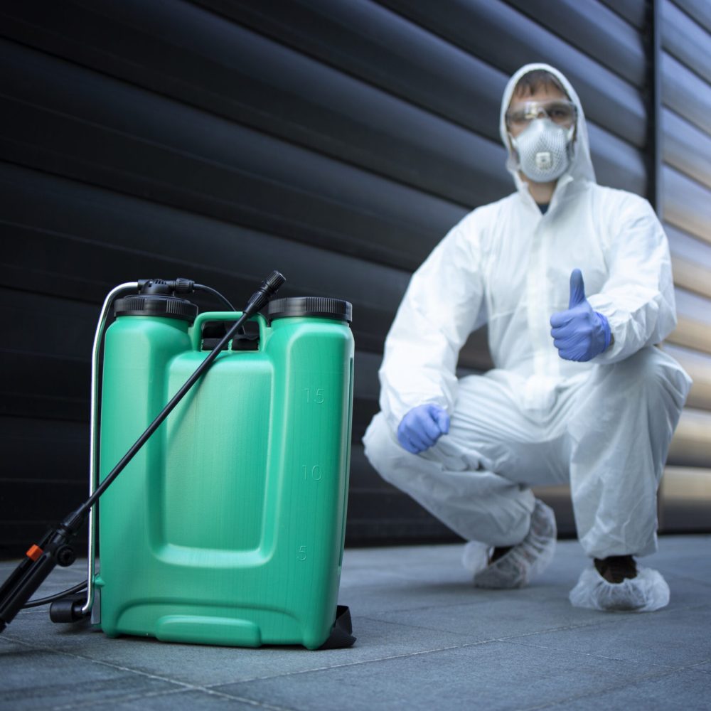 Exterminator in white protective uniform standing by reservoir with chemicals and sprayer.